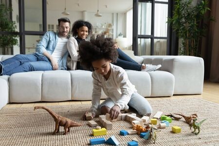 African daughter play on floor with wooden blocks set and dinosaur toys while multi ethnic parents sitting on sofa in living room, homeowners spend time at new home. First house family weekend conceptの写真素材