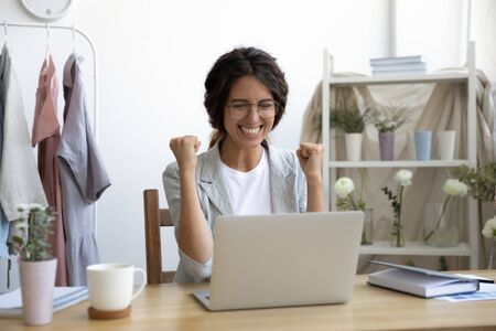 Happy young Caucasian woman in glasses sit at desk look at laptop screen triumph receive good news in email, excited female feel overjoyed euphoric win online lottery on computer, luck conceptの写真素材