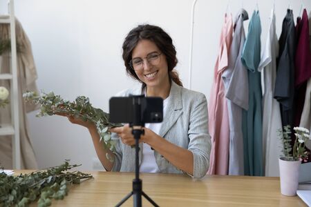 Smiling young Caucasian female florist tutor shoot video blog of floral compositions design on cellphone camera, happy woman designer record vlog tutorial on cell about handmade bouquet home decorの写真素材