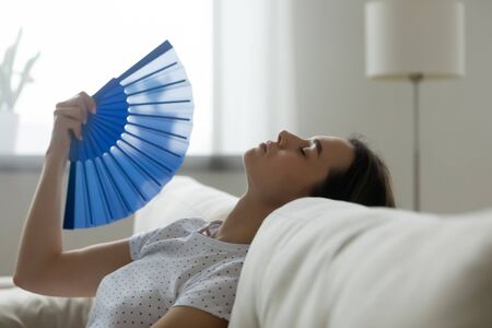 Close up image woman puts head on sofa cushions closed eyes feels sluggish due unbearable heat, waves hand blue fan cool herself, hot summer flat without air-conditioner climate control system conceptの写真素材