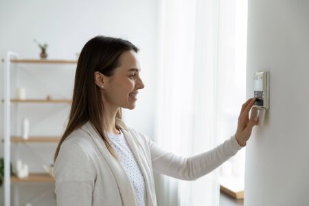 Smiling woman standing near wall-mounted device adjusting degrees in living room set comfortable temperature using thermostat home heating system. Owner of modern smart house, energy saving conceptの写真素材