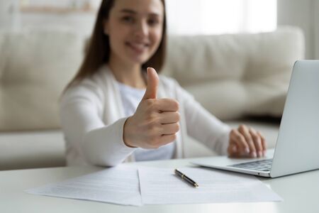 Woman sit at coffee table at home near laptop smile show at camera thumbs up gesture, close up. Good terms new mobile e-bank, e-services, buy insurance, satisfied client give positive feedback conceptの写真素材