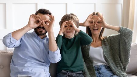 Head shot portrait happy parent with little son making funny faces, binoculars glasses eyewear shape gesture, looking at camera through fingers, sitting on cozy couch, family having fun togetherの写真素材