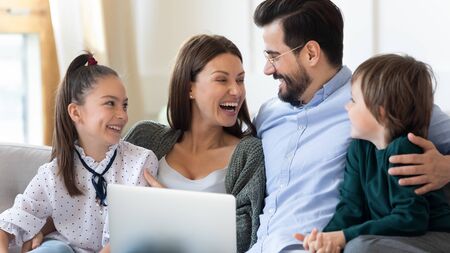 Laughing mother and father with children using laptop at home, sitting on couch, family having fun together, excited parents with little son and daughter shopping or watching funny video onlineの写真素材