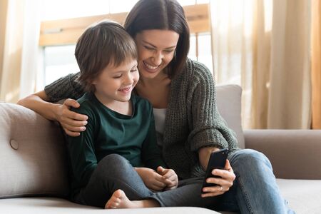 Happy mother and little son using smartphone together, sitting on sofa, looking at phone screen,...
