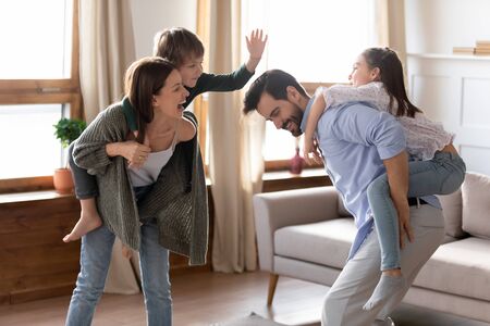 Happy parents carrying children on backs and running, family having fun at home, excited mother and father playing with adorable little son and daughter in modern living room, piggy backの写真素材