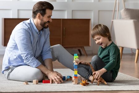 Happy father and little son playing with toys together, laughing, having fun, sitting on warm floor with underfloor heating, smiling dad and cute boy, building tower from colorful wooden blocksの写真素材