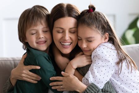 Head shot portrait happy mother hugging little son and daughter with closed eyes, smiling beautiful mum and adorable children cuddling, enjoying tender moment together, expressing love and careの写真素材
