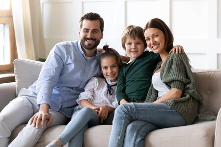 Family portrait happy parents with cute little son and daughter sitting on cozy couch at home, posing for photo together, looking at camera, smiling beautiful mother and father hugging kidsの写真素材
