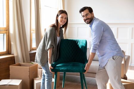 Portrait happy young couple carrying chair, placing furniture in living room in new house, looking at camera, moving day and renovation concept, excited family relocating into new apartmentの写真素材