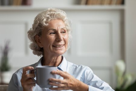 Attractive happy old woman holds coffee tea cup enjoy hot beverage resting on sofa in cozy living room at home. Granny looking at distance feels healthy and optimistic, relish life and new day conceptの写真素材