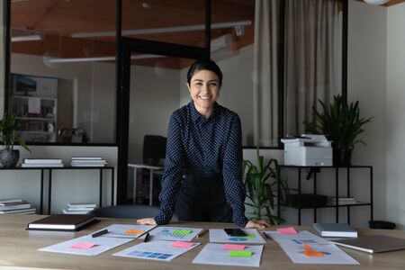Portrait of successful talented project manager. Leaned her hands on desk Indian ceo smile looks at camera feels prepared for presentation, printed data lie at table for clients, or staff for seminarの写真素材