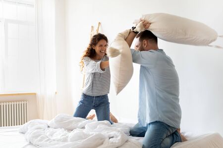 Happy young couple pillows fight and laughing on bed at bedroom. Smiling bearded man and woman...