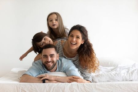 Happy young parents with little children lying on bed looking at camera. Smiling beautiful mother and bearded father enjoying free time with cute adorable daughter and son.の写真素材