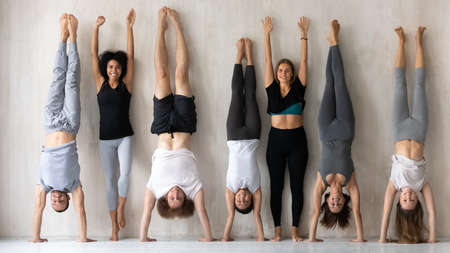 Happy young multiracial women in activewear standing near wall with raised arms while sportive mixed race people performing handstands, having fun together before intensive workout in gym indoors.の写真素材