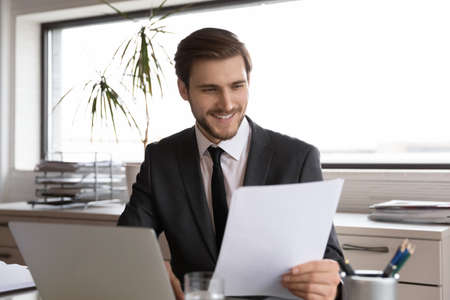 Smiling young Caucasian male employee sit at desk in office work on laptop consider company paperwork, happy businessman use computer read good rates numbers on financial document or reportの写真素材