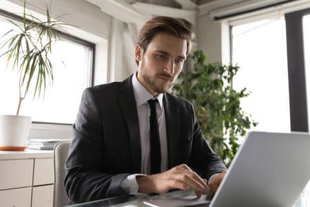 Concentrated Caucasian male employee sit at desk in office work on laptop consult client or customer online, focused young businessman busy using computer internet connection, technology conceptの写真素材