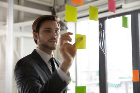 Focused young male employee write ideas on sticky notes on glass wall in office, engaged in creative thinking, motivated businessman develop company startup plan or project, brainstorm in boardroomの写真素材