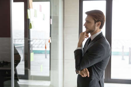 Pensive young Caucasian businessman look at glass wall in office think of project implementation, thoughtful serious male employee brainstorm develop business plan in boardroom, work with sticky notesの写真素材