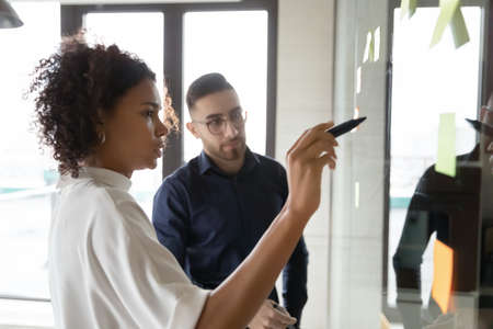 Focused multiracial employees write ideas on glass wall plan startup in office together, diverse...