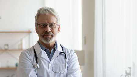Headshot portrait of serious mature male doctor or therapist in white medical uniform, glasses and stethoscope, mature old man GP or physician in spectacles pose at workplace, healthcare conceptの写真素材
