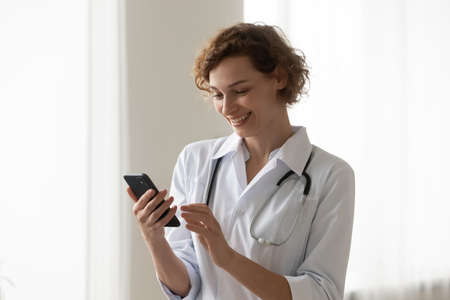 Smiling Caucasian young female nurse browse internet on modern cellphone gadget in clinic, happy...