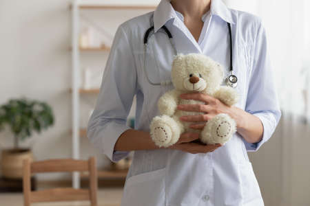 Crop close up of female medical specialist or nurse in white uniform hold stuffed teddy bear in clinic, woman doctor or GP with plush toy show care and love to little patient children in hospitalの写真素材