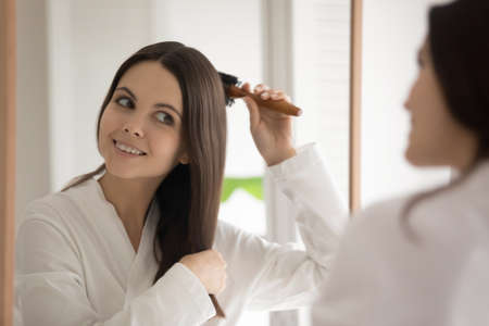 Mirror reflection head shot smiling beautiful young woman combing hair with wooden brush after shower, satisfied attractive girl wearing white bathrobe standing in bathroom, enjoying morning routineの写真素材