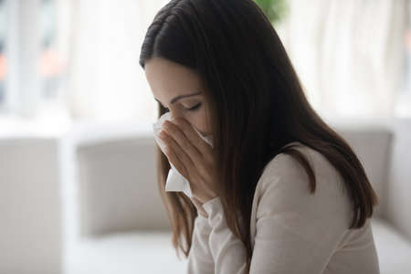 Close up sick exhausted young woman blowing nose, holding using white paper napkin handkerchief, symptom of chronic sinusitis, cold or flu, seasonal allergy, unhealthy girl feeling unwellの写真素材