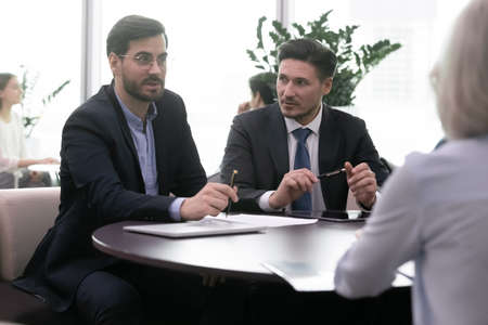 Serious business partners working together on corporate meeting. Two businessmen sit in front of businesswoman and cooperate. Successful entrepreneur clients consult with expert or explain dealの写真素材