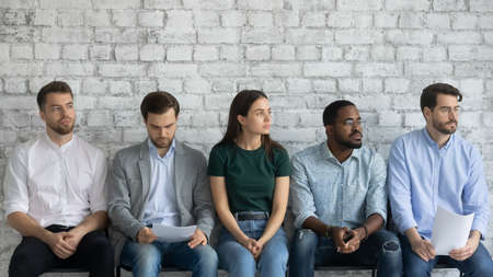 Five bored multiracial unemployed applicants sit in line against brick wall background looking away...