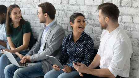 Cheerful diverse applicants hold devices talking while waiting turn job interview. People gather in office prepared for seminar, communicating enjoy chat and friendly relations in collective conceptの写真素材