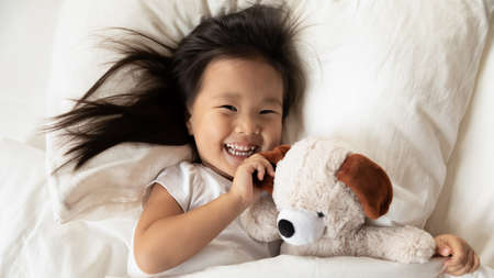 Happy small adorable vietnamese kid girl lying on pillow under duvet, looking at camera, feeling energetic after waking up in morning, playing with toy staying in bed, above top head shot close up.の写真素材