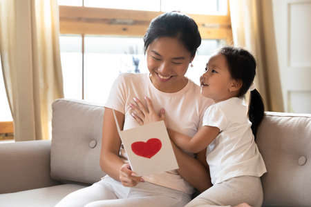 Grateful smiling young asian ethnic mother looking in handmade paper card, watching funny picture from preschool cute kid daughter, enjoying special occasion birthday wishes, feeling happiness.の写真素材