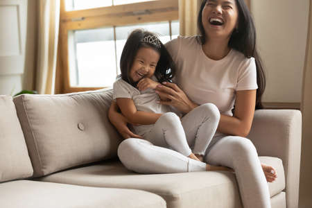 Overjoyed young vietnamese ethnic woman mother sitting on cozy couch, tickling little adorable funny preschool asian kid daughter, enjoying playtime together at home, laughing joking indoors.の写真素材