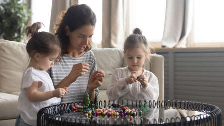 Caring young mother and two adorable little daughters making colorful beads jewelry together, sitting at table in living room, happy mum and cute preschool girls enjoying creative activityの写真素材