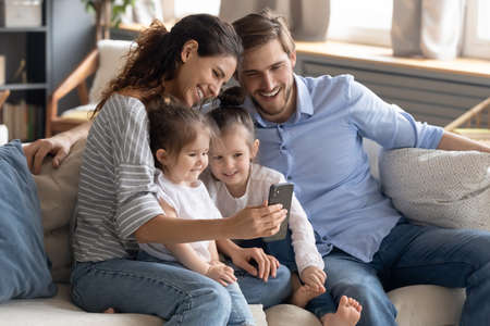 Smiling young mother and father with two adorable little daughters using smartphone, looking at...
