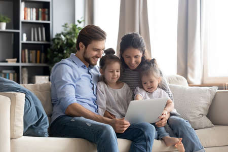 Happy young family with two little daughters using laptop together, smiling mother and father with adorable preschool girls looking at screen, watching cartoons, shopping online, making video callの写真素材
