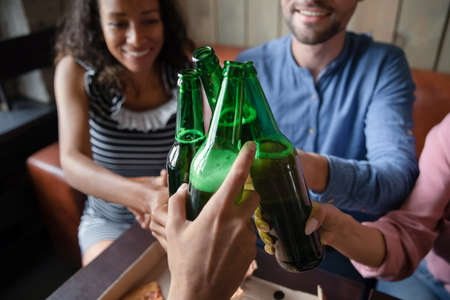 Group of multi racial best friends clinking glass bottles of beer while celebrating life event sit in bar public place gather together enjoy time with mates drink alcohol close up, hanging out conceptの写真素材