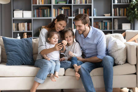 Happy young parents with two adorable little daughters using phone, looking at screen, sitting on...