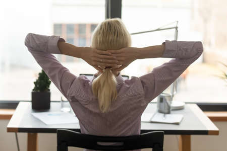Back view of exhausted blonde woman lean in chair relax at workplace, take nap or daydream, tired female employee rest at table, breathe fresh air, relieve negative emotion, stress free conceptの写真素材