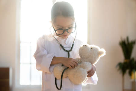 Close up pretty little girl playing doctor, using stethoscope, holding fluffy toy bear, checking heart or breath, preschool child wearing white coat uniform and glasses pretending pediatricianの写真素材