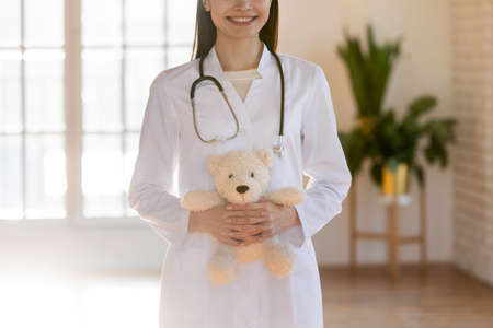 Close up smiling doctor pediatrician holding fluffy toy teddy bear in hands, standing in modern clinic cabinet, therapist gp wearing white coat uniform with stethoscope, childcare medical serviceの写真素材
