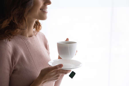 Crop close up of smiling young Caucasian woman hold cup drinking tasty black aroma tea at home,...