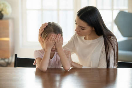 Caring mother calming and hugging crying upset little daughter, sitting at desk together, loving mum expressing support, comforting offended preschool girl, children psychologist conceptの写真素材