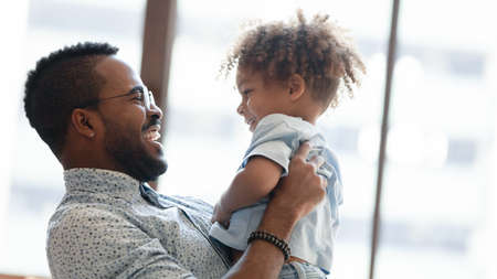 Close up of smiling young biracial dad hug cuddle cute little ethnic son playing together at home,...