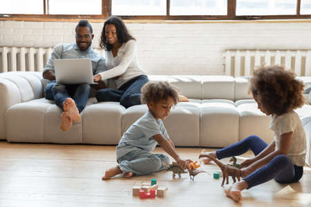 Little biracial brother and sister children sit on warm wooden floor in living room play toys together, happy african American parents couple relax on couch using laptop, ethnic family enjoy weekendの写真素材