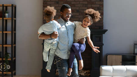 Happy young biracial father hold in arms play with two little kids at home, enjoying weekend together, smiling african american dad have fun engaged in funny activity game with excited small childrenの写真素材
