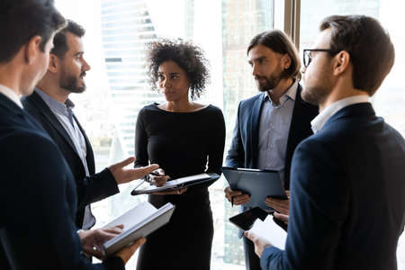Focused young multiracial diverse business people in formal wear standing in circle, holding clipboard with paper reports, sharing project ideas opinions at informal conversation in modern office.の写真素材