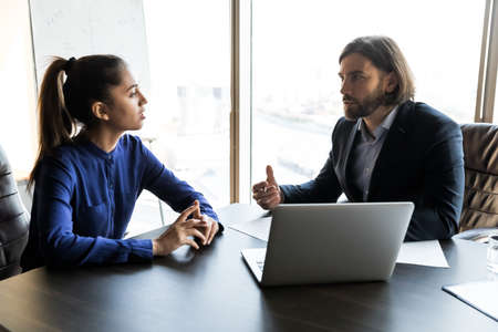 Serious young male boss in formal wear discussing working issues with millennial motivated female...